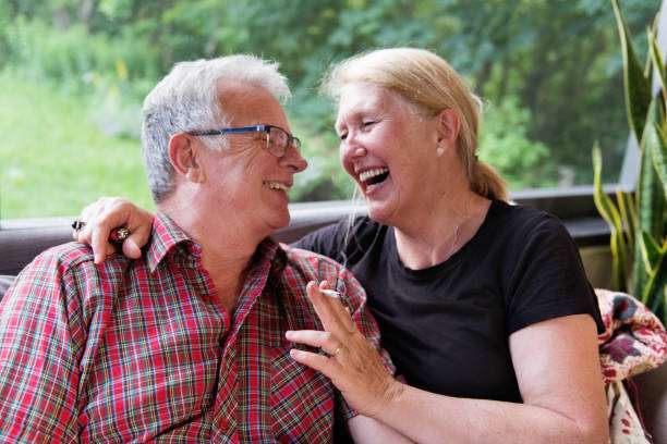 Senior retired couple enjoying marijuana in their gazebo garden. They smoke medicinal cannabis. They have 15 years of age difference. She is 60 years old and her husband is 72 years old. The women have blond hair and the men have gray hair. She wear pants and t-shirt and he is wearing shorts and skirt. It is summer and the photo was taken in Quebec Canada.