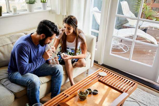 Smiling young couple sitting on their living room sofa at home and smoking marijuana in a bong