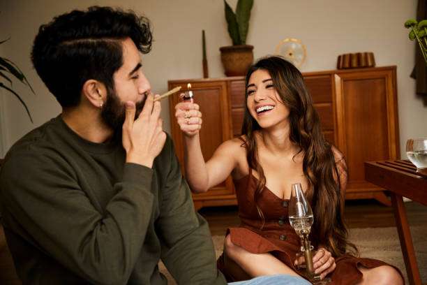Laughing young woman lighting a joint for her boyfriend while getting high together in their living room