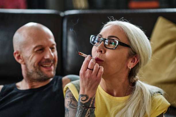 Selective focus shot of mature Caucasian woman with long gray hair and tattoos on arm smoking cannabis cigarette